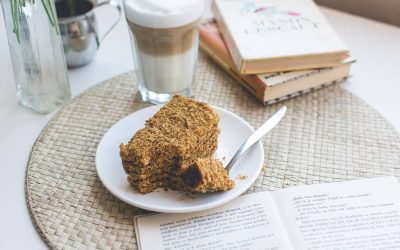 A piece of cake on a plate next to a book and a cup of coffee.